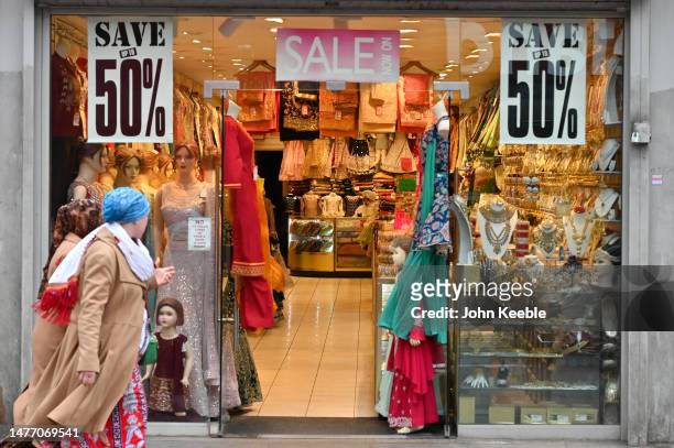 People walk past a fashion shop advertising a 50% sale selling sarees, lehengas and bridal jewellery in Upton Park Green Street on March 23, 2023 in...