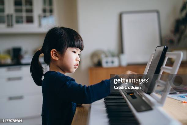 little asian girl playing piano in the living room at home, using a digital tablet for sheet music. hobbies and interests - keyboard musical instrument child stock pictures, royalty-free photos & images