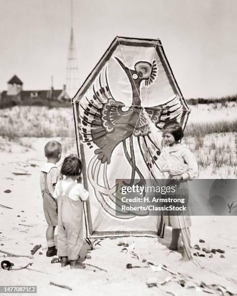 1920s girl on beach with large kite painted with a big bird a phoenix talking to two younger kids boys in awe of kite's size.