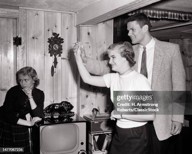 1950s teen boy giving teenage girl hands on help aiming a dart during party in knotty pine rec room another girl leaning on tv.