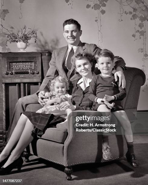 1940s happy smiling family of four sitting together in living room chair by console radio dad mom son daughter looking at camera.