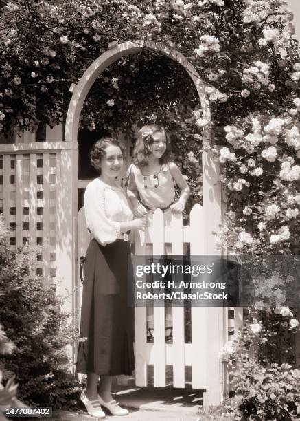 1930s smiling woman mother and girl daughter standing by white picket fence garden gate in surrounding blossoming rose arbor.