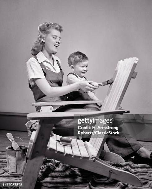 1940s 1950s smiling woman mother painting adirondack chair with help from boy her laughing toddler son.