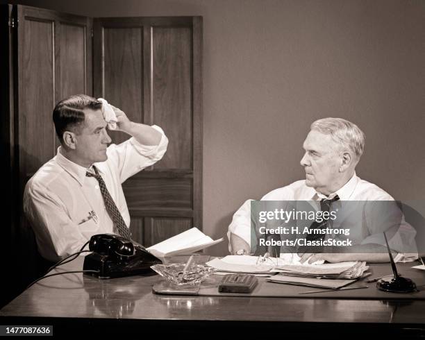 1950s two men at desk rolled up sleeves working with papers one man sweating older man with stern facial expression.