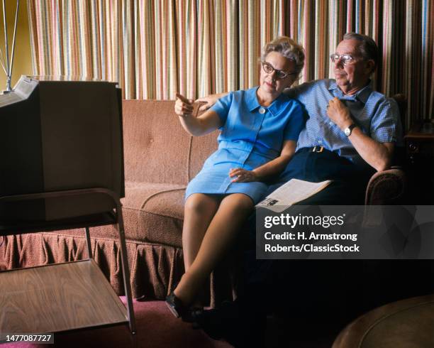 1970s couple sitting on couch side by side watching a portable television in the evening.