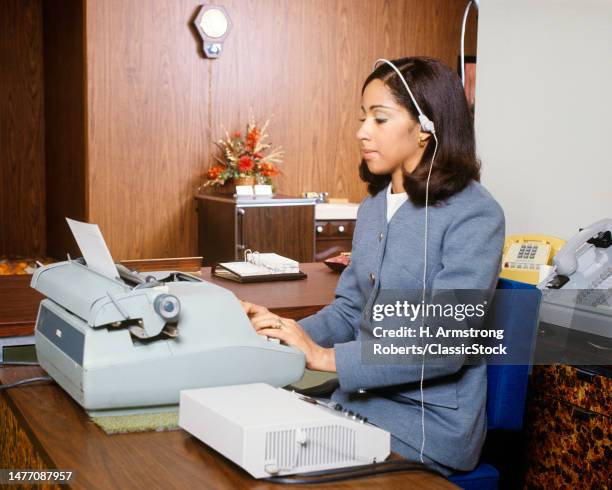 1970s woman in office typing on electric typewriter listening to dictation from dictaphone with headset.