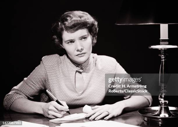 1940s 1950s serious thoughtful married brunette woman with pen in hand about to write a check she is wearing a polka dot blouse.
