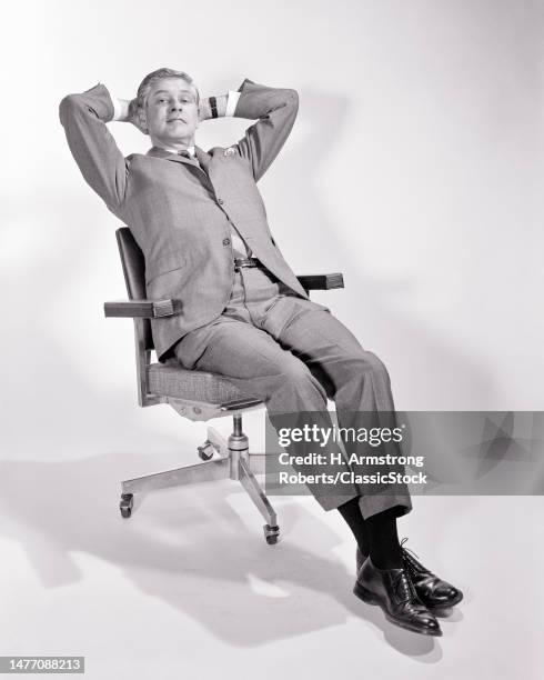 1960s businessman sitting in office chair leaning back hands behind his head looking at camera.