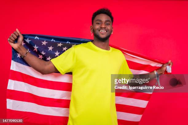 young african american man smiling with usa flag, cheerful black man looking at camera proudly isolated on red background, celebrating independence day, 4th of july - classic day 4 stock pictures, royalty-free photos & images
