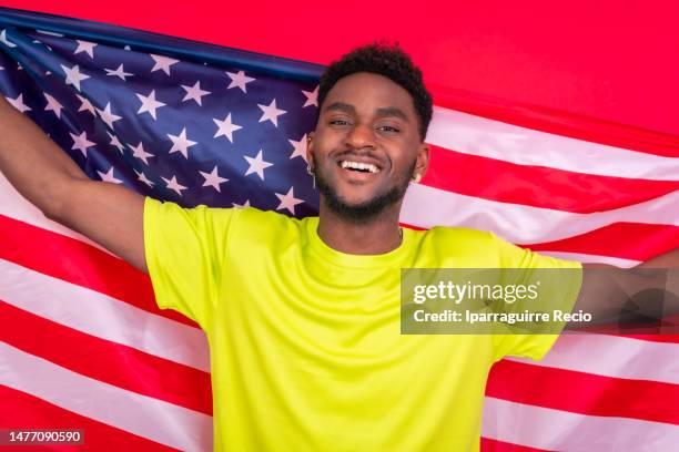 young african american man smiling and holding up usa flag, cheerful black man looking at camera proudly isolated on red background, celebrating independence day, 4th of july - classic day 4 stock pictures, royalty-free photos & images