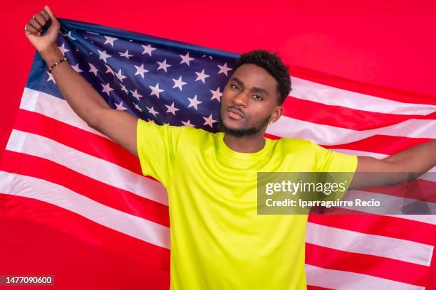 young african american man smiling and holding up usa flag on his back, cheerful black man looking at camera proudly isolated on red background, celebrating independence day, 4th of july - classic day 4 stock pictures, royalty-free photos & images