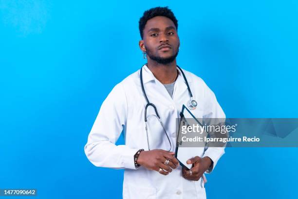 portrait of young black ethnicity, doctor or nurse in blue gown and stethoscope, with medical file folder, isolated on blue studio background - man standing full length stock pictures, royalty-free photos & images