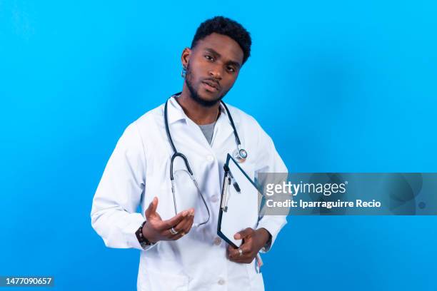 portrait of young black ethnicity man with serious look, doctor or nurse in blue gown and stethoscope, with medical file folder, isolated on blue studio background - man standing full length stock pictures, royalty-free photos & images