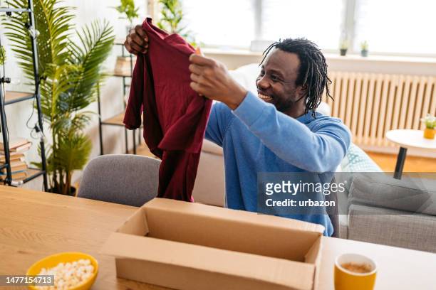 junger mann packt ein paket mit einem neuen hemd aus, das er online bestellt hat - online shopping opening package stock-fotos und bilder