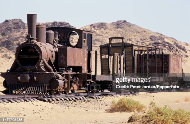 The rusting remains of a narrow gauge steam locomotive and trucks on rails in a siding of the Ottoman Empire era Hejaz Railway at the abandoned...