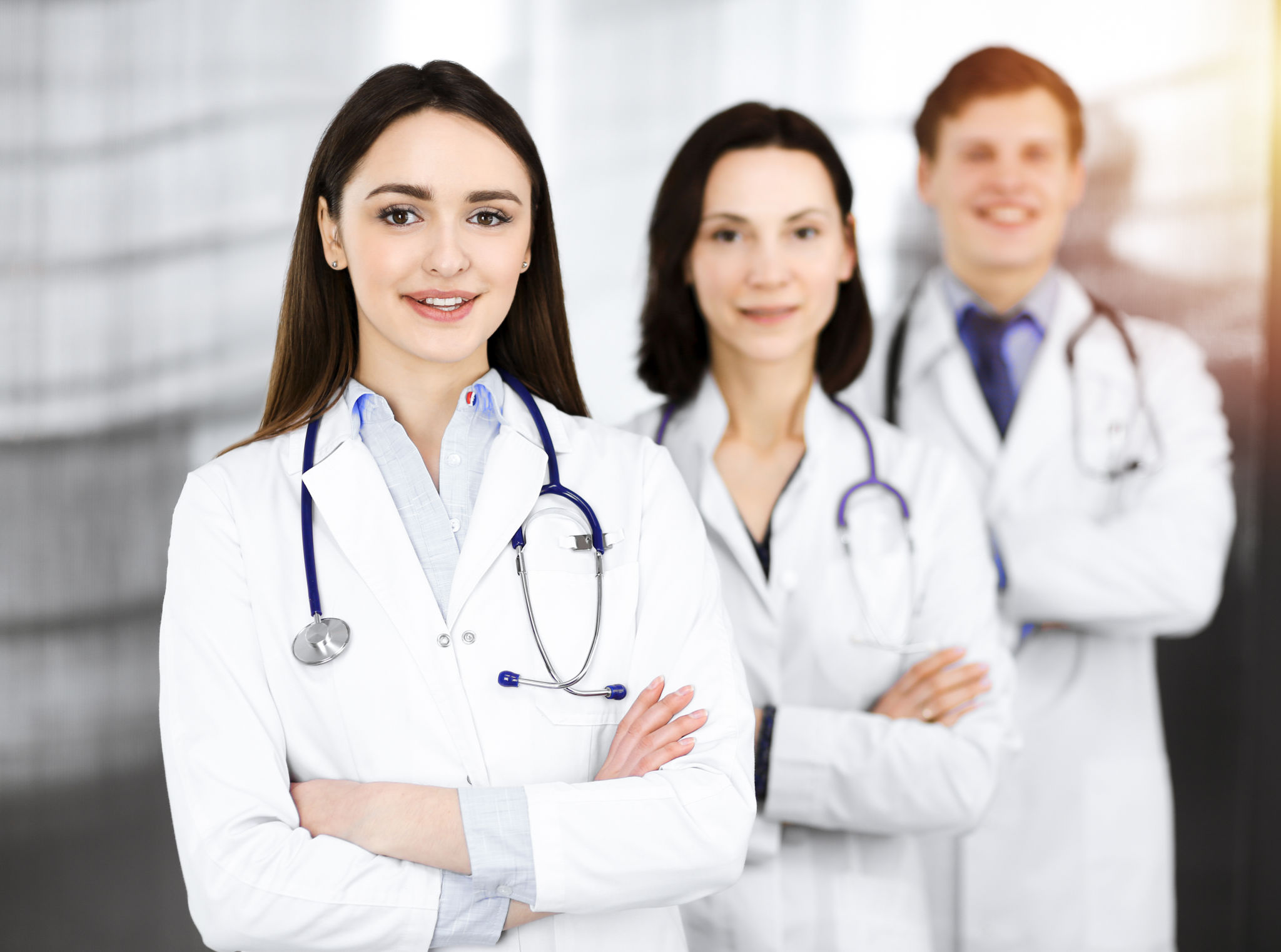 Group of young professional doctors standing as a team with arms crossed in sunny hospital. Medical help, insurance in health care, best disease treatment and medicine concept Group of young professional doctors standing as a team with arms crossed in sunny hospital. Medical help, insurance in health care, best disease treatment and medicine concept