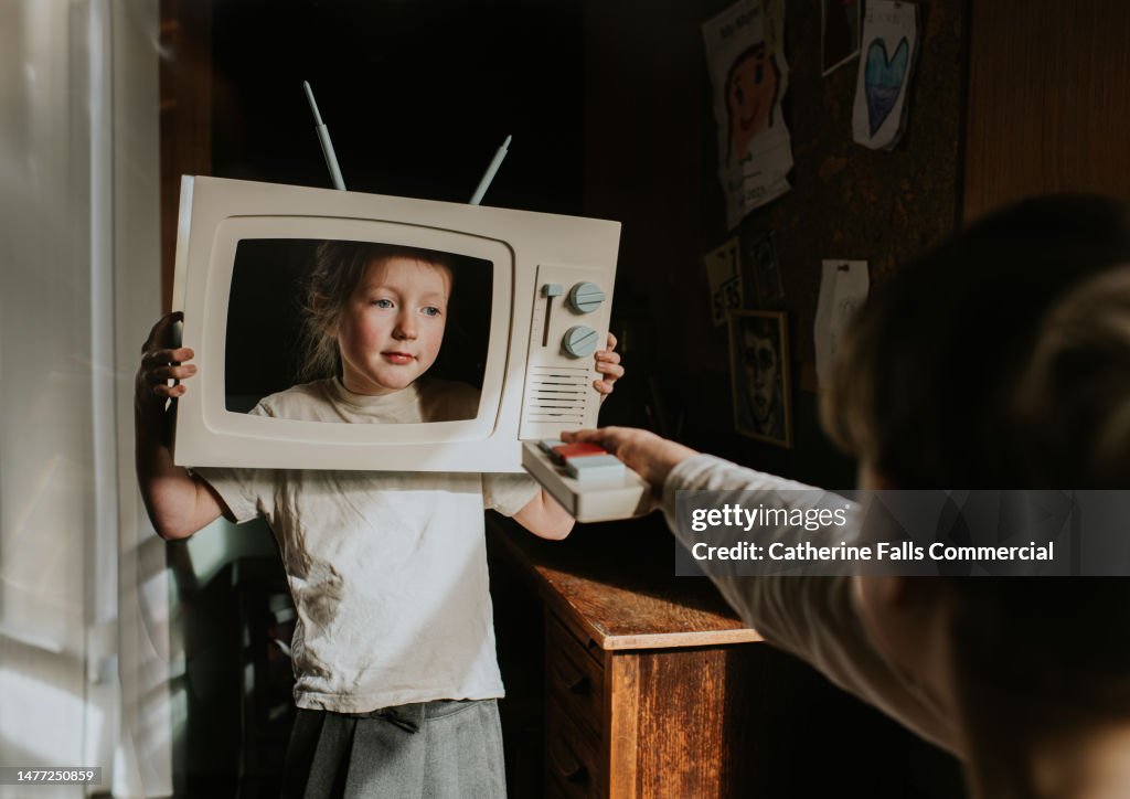 A child pretends to be the star of her own TV show, while her brother holds the remote up to change the channel