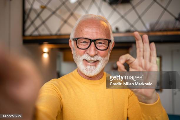 lächelnder älterer mann, der zu hause im wohnzimmer ein selfie macht - one finger selfie stock-fotos und bilder