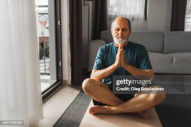 elderly man with beard praying and meditating at home, practicing yoga in lotus position - man meditating stock pictures, royalty-free photos & images