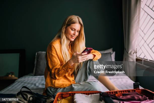 a happy beautiful blonde businesswoman texting on her mobile phone while packing her suitcase for a business trip - woman packing suitcase stock pictures, royalty-free photos & images