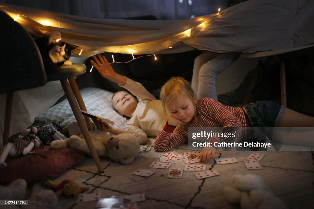 A brother and a sister playing with their cat in a homemade illuminated hut, at home
