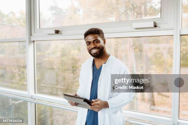 portrait of smiling young male doctor holding digital tablet standing against window at hospital corridor - young adult at doctor stock pictures, royalty-free photos & images