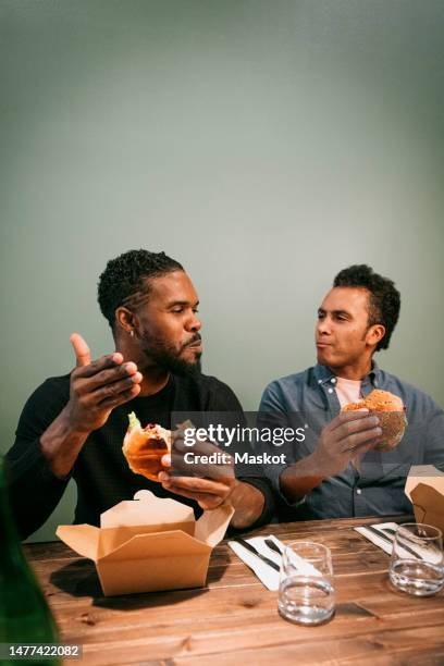 male friends talking to each other while eating burgers sitting at restaurant - male burger eating fotografías e imágenes de stock