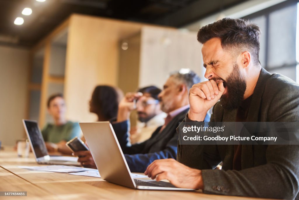 Young businessman yawning while working on a laptop in a busy office lounge