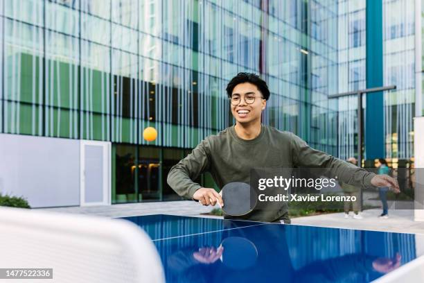 happy young asian man having fun playing table tennis with colleague in an office zone. chinese young male enjoying ping pong gam with friend. leisure activity concept. - male colleague stock pictures, royalty-free photos & images