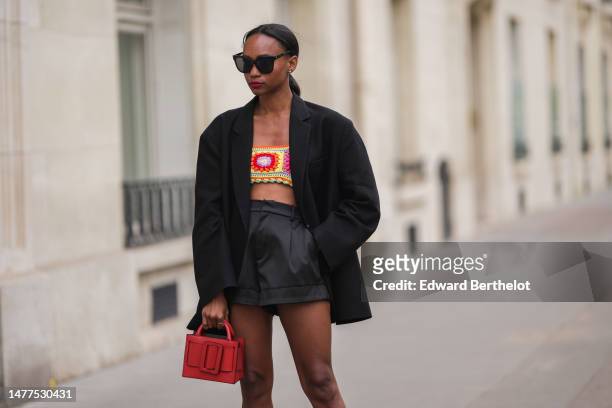 Emilie Joseph wears sunglasses, a black oversized blazer jacket by The Frankie Shop, black high waist satin shorts, a colored yellow and red crochet...