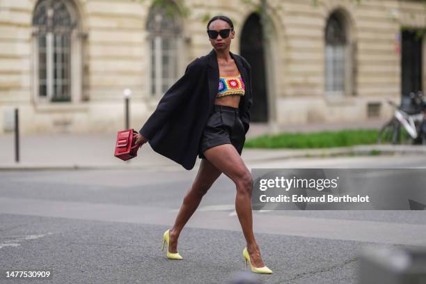 Emilie Joseph wears sunglasses, a black oversized blazer jacket by The Frankie Shop, black high waist satin shorts, bright pastel yellow leather...