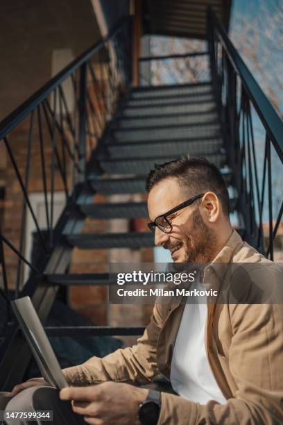 a young man sits on the steps in front of his house and uses a laptop for online work or surfing the internet - mid volwassen vrouw stockfoto's en -beelden