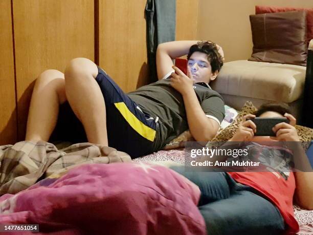 two boys busy in using their mobile phones in a living room while lying on their backs. - teen boy shorts stock pictures, royalty-free photos & images