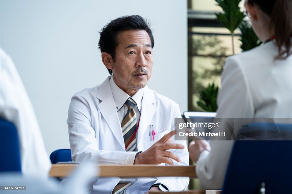A doctor man explains to a pharmaceutical salesman and his colleagues in a conference room.