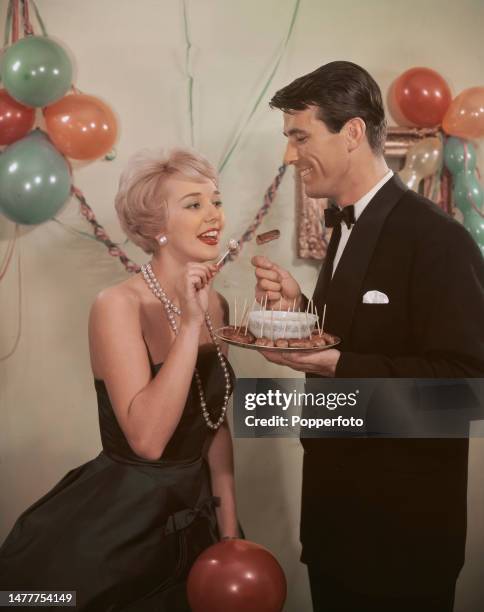 Posed studio portrait of a house party scene with a man and a woman eating cocktail sausages on sticks with a dipping sauce, he wears an evening...