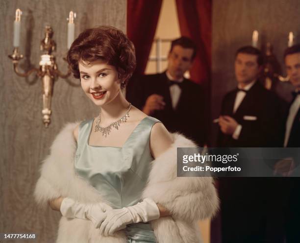 Three men in evening wear attending a social function look towards a woman posed wearing a silver satin evening dress, white gloves and white fur...