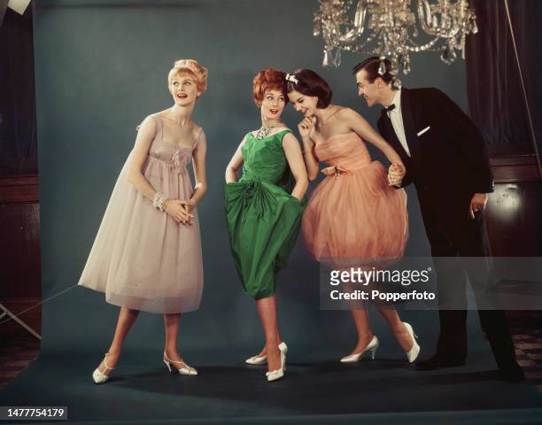 Posed studio portrait of a man in eveningwear with three female fashion models wearing, from left, an empire line ballgown in pale pink chiffon, a...