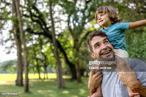 padre feliz cargando a su hijo sobre sus hombros en el parque - llevar al hombro fotografías e imágenes de stock