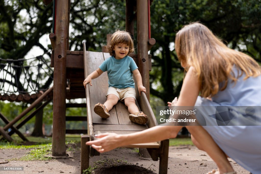Junge spielt im Park auf der Rutsche unter Aufsicht seiner Mutter
