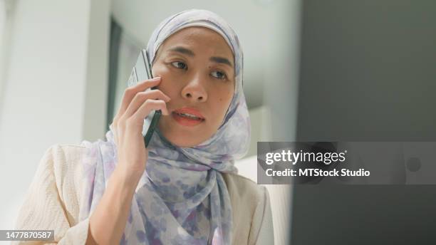 closeup of happy young muslim woman sit on bed with computer laptop and using mobile phone talk with colleague meeting work in bedroom at home. work from home. - worry business stock pictures, royalty-free photos & images