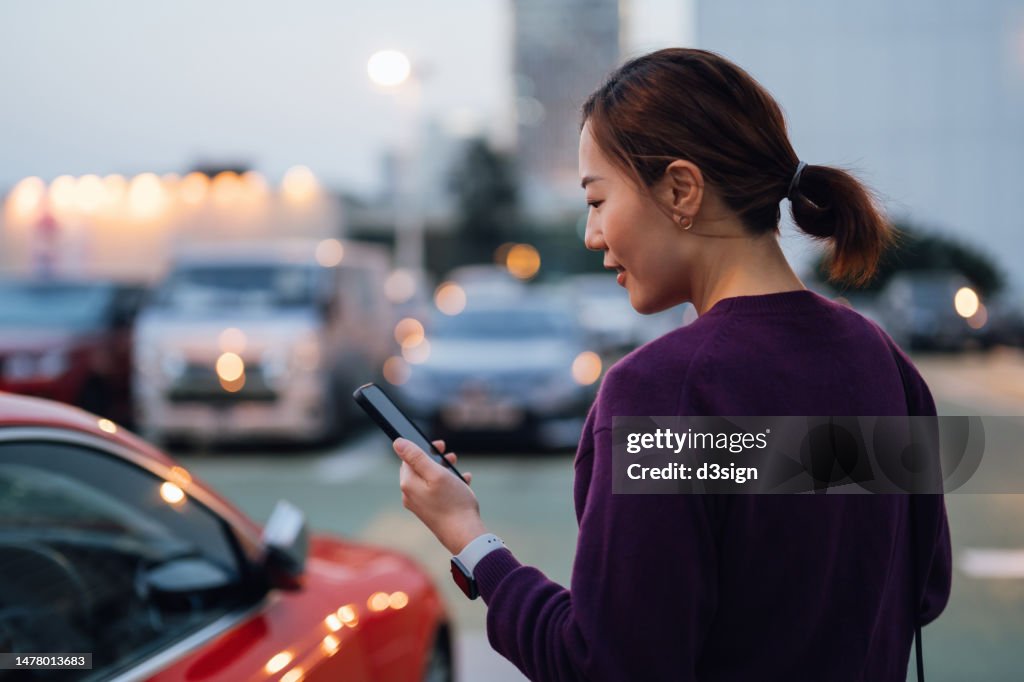 Young Asian businesswoman using smartphone while walking to her car, commuting in the city, with urban cityscape in background. Business on the go concept