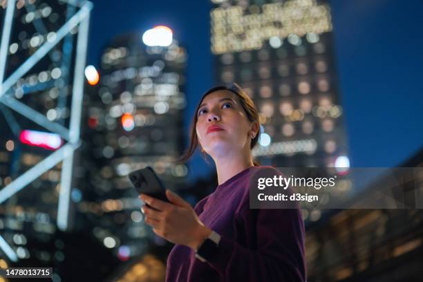 low angle portrait of confident and determined young asian businesswoman using smartphone while standing against illuminated corporate skyscrapers in financial district in the city. female leadership and empowerment. business success and commitment - low confidence stock pictures, royalty-free photos & images