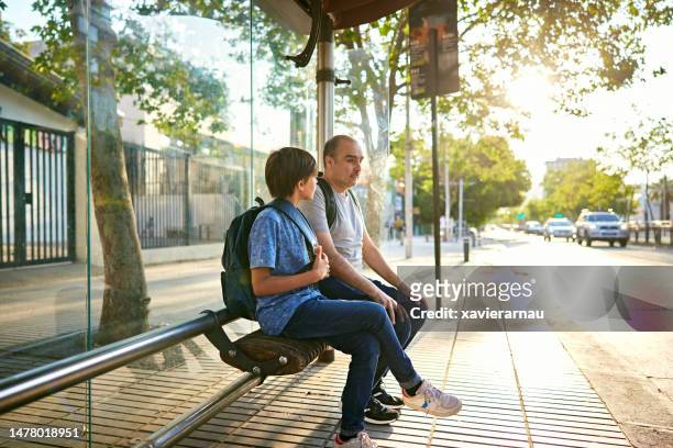 boy and his father waiting for transportation at bus shelter - bus shelter stock pictures, royalty-free photos & images