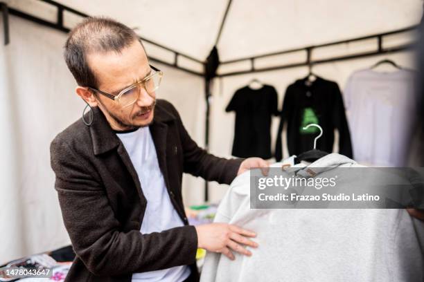 mature market vendor organizing his clothes for sale at market stall - entertainment tent stockfoto's en -beelden