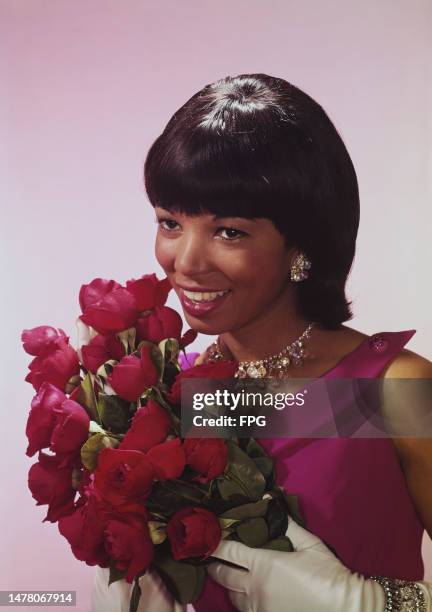 Woman, wearing a sleeveless fuchsia pink outfit with white evening gloves and a silver necklace, silver bracelet and silver earrings, smiles as she...