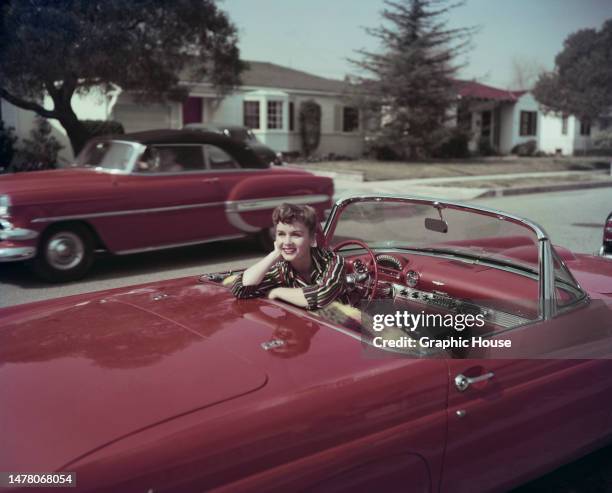 American actress and singer Debbie Reynolds smiles while sitting in the driver's seat of a red convertible Ford Thunderbird, turning in her seat to...