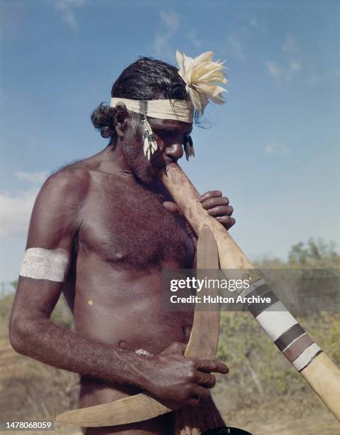 An Australian aborigine wearing a headband adorned with a flower as he holds a boomerang while playing the didgeridoo, Australia, circa 1965.