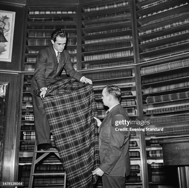 Shop assistant standing on a step ladder at a Scotch Wool Shop, shows a length of tartan fabric to another man, London, December 4th 1958. Behind...