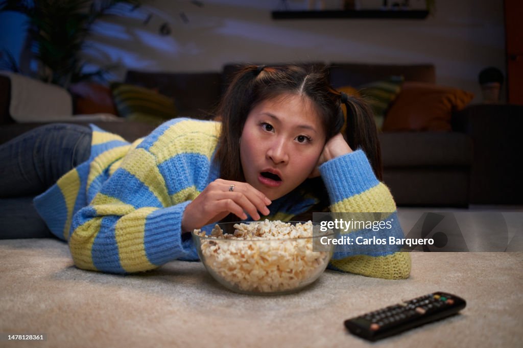 Young Asian girl lying on the living room floor watching a movie at home, amazed by what's on TV.