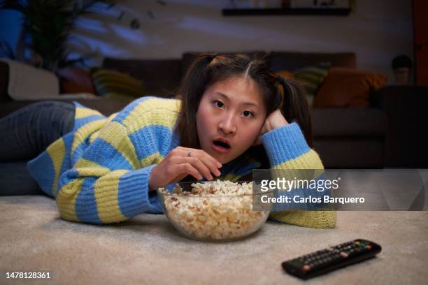 young asian girl lying on the living room floor watching a movie at home, amazed by what's on tv. - epic film stock-fotos und bilder